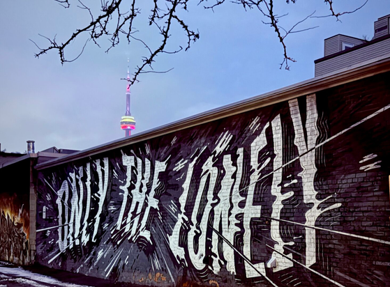 Low rise brick building painted with ONLY THE LONELY in white letters along a wavy line. Background is grey/black with lines coming from behind THE, warping and stretching at the letters outwards in a starburst. Background is clear sky at dusk, the top of the CN Tower is visible.