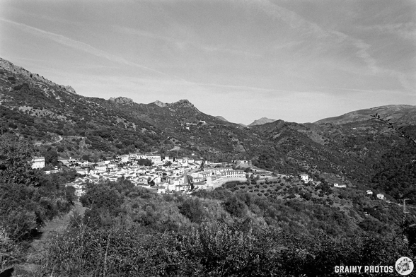 A panoramic view of a mountainous landscape featuring Benadalid nestled among rolling hills and greenery, captured in black and white. The scene highlights the natural beauty and serene atmosphere of the area.
