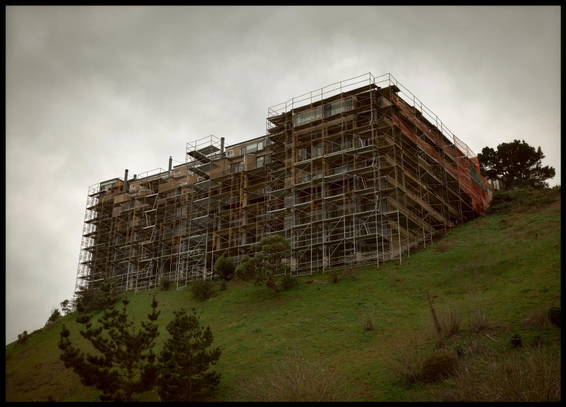 A tall building rises like the prow of a battleship over a steep slope in the Diamond Heights, Goldmine Hill neighborhood. The building is surrounded by scaffolding as work proceeds on the exterior siding. 

The photo was taken on a dark afternoon in the rainy season (winter here). So there are gray skies above and green grass on the slope, with a few bushy things clinging to the otherwise sparsely vegetated earth.
