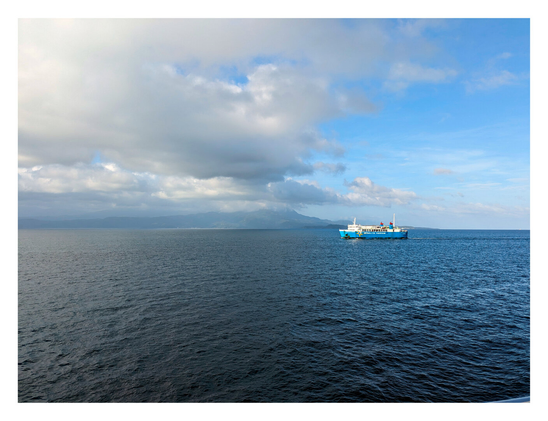 A blue and white ferry boat traveling across deep blue waters. In the distance, a hazy, mountainous landmass sits along the horizon. Above, the sky is split between large, billowing white clouds on the left and clear blue sky on the right. - Google Gemini 3 Pro Preview