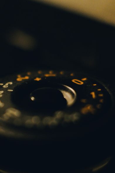 Macro photograph of the exposure compensation dial on a camera. Half the dial is covered in red indexes going from -2 to +2 in one-third steps, and the other half has indexes going from -3 to +3 in half steps. In the middle of the dial sits a rounded push-down button to lock the dial's rotation. Everything else is a dark and nondescript blur.