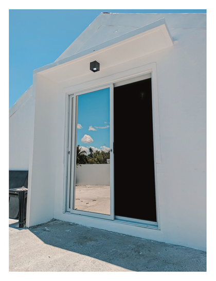 A slid sliding glass door that leads to the second floor of the house. Sky, clouds, and trees can be seen in the reflection of the door. Above the door is a modern outdoor ceiling light on the overhang.
