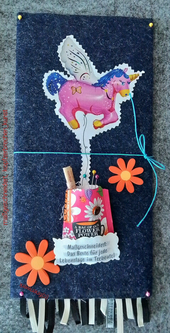 mixed Media arbeit; auf einer dunkelblauen Filzunterlage ein Bild eines rosa EINHORN Luftbalons, dessen Schnur in einem Teebeutel endet. Nähzeug befindet sich im geöffneten Beutel. Ein Schild weist  den Bildtitel: maßgeschneidert: das Beste für jede Lebenslage im Teebeutel. Zwei orangene Dekoblüten,  und schwarz weiße Gummifranzen runden das Ensembles ab.