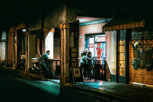 In a dimly lit scene, patrons gather outside a pizzeria at night. Most of the photo dominated by dark shadows. Two people are seated at a small table just inside the door of the cafe with a worker right behind them. There are neon signs and posters are visible through the window behind them. To the left of the cafe entrance, a man stands alone at a high table, looking at his phone. He is dressed casually in a t-shirt and shorts. To the right of the cafe entrance, the metal security shutters are pulled down and are covered in graffiti.