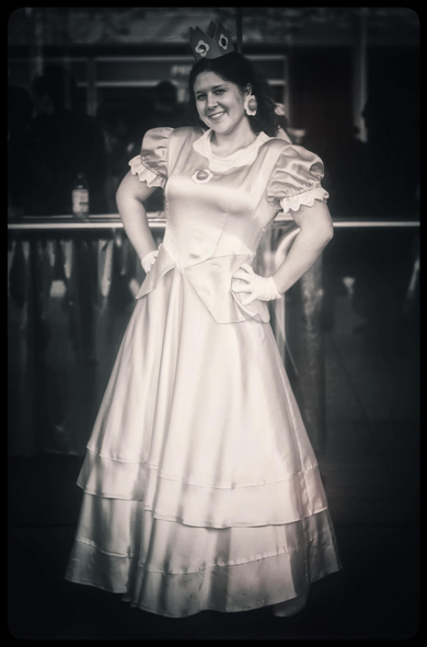 A monochrome portrait of a young lady in a silvery, Disney-esque long dress with puffed shoulders, standing tall with hands on hips as if posing for a movie poster. She has white gloves, a big smile, large floral earrings, and a small crown on top of her head. 

She looks radiant and rather magical, glowing against the dark background of the convention hall.
