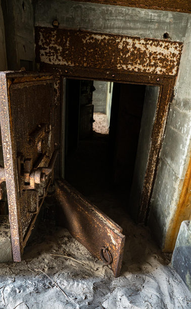 Color photo of a large and heavily rusted open steel door and sand-covered floor below it revealing the entry to a dark, underground military bunker from the Nazi occupation of Denmark during WWII. 