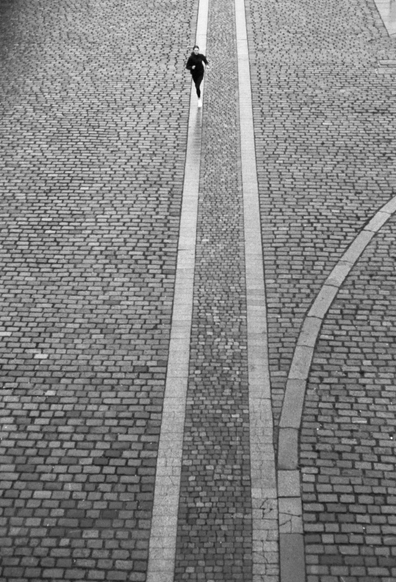 A person is running along a cobblestone street. The scene is captured from above, showcasing two distinct pathways marked by lighter stones. The image is in black and white.
