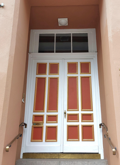 Entrance to a façade in light apricot. Four steps lead up to the door, flanked on the left and right by brass-coloured railings. Double-winged panel door in classicist style with white and gold frames, panels in the colour of ripe apricots. Above it is a three-part glass skylight and a white, modern lamp.  An ensemble reflecting the proud and self-confident attitude of Lübeck's upper middle class at that time.