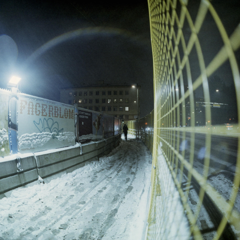 City scene at night during winter. Fencing along a building site, someone walking between the fences, hunching down form the snowfall.