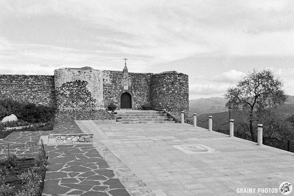 A historic stone fortress, now a cemetery, stands on a rocky terrain, featuring a large entrance door and surrounded by a scenic landscape. The structure is partially surrounded by trees and offers a panoramic view of the mountains in the distance, captured in black and white.