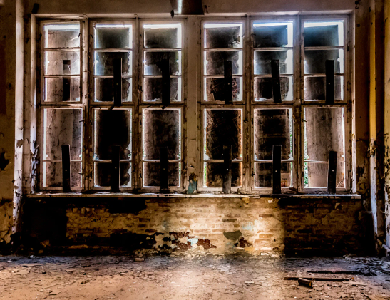 Fensterfront in einem verlassenem Gebäude. Sechs schmale, hohe mehrteilige Fenster. Alle sind mit Sperrholz vernagelt, dass an den Rändern noch Licht herein lässt. 
Mittig in den Raum ein Lichtschein, der von der geöffneten Tür kommt.