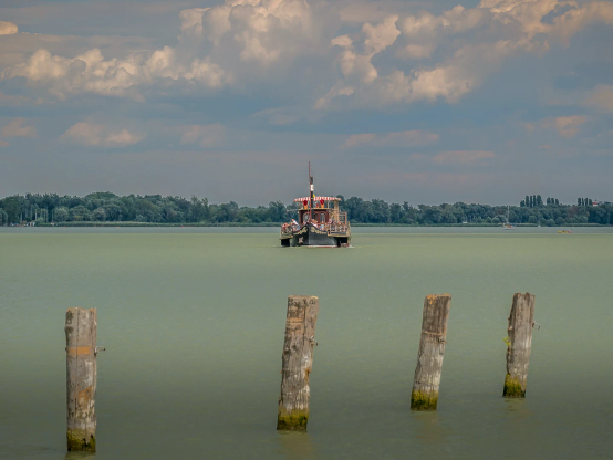 Vor grauem Himmel präsentiert sich der Balaton, der heute meergrün trägt. Im Vordergrund vier Holzpflöcke in Reihe, die im Wasser stehen. Ein Ausflugsboot im alten Stil hält auf uns zu. 