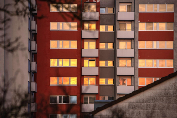 Das Bild zeigt die Fassade eines großen, mehrstöckigen Wohngebäudes bei Sonnenuntergang, das aus mehreren vertikalen Abschnitten besteht. Der linke und rechte Abschnitt sind in einem kräftigen Rot gestrichen, während der mittlere Abschnitt grau ist. Alle Fenster in den roten Abschnitten spiegeln das intensive, warme, orange-gelbe Licht der tief stehenden Sonne wider, was einen starken Kontrast zur dunkleren Umgebung bildet. Die Fenster in den grauen Abschnitten zeigen ebenfalls dieses warme Licht, jedoch in geringerem Maße.
Jeder Abschnitt hat mehrere Etagen mit Balkonen, die durch weiße Brüstungen gekennzeichnet sind. Im Vordergrund, im unteren rechten Bereich, ist das schräge, dunkel gedeckte Dach eines kleineren, helleren Gebäudes teilweise sichtbar. Am linken und oberen Rand des Bildes verdecken die dunklen, unscharfen Umrisse von Baumzweigen teilweise die Gebäudestruktur, was dem Bild Tiefe verleiht.