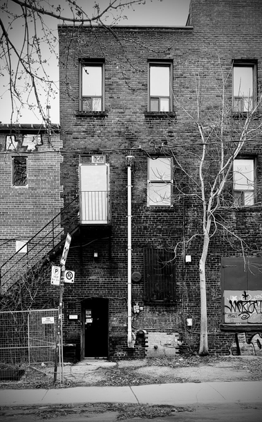 Brick façade of a 3-storey building. Industrial-style metal door on the first floor, with 2 windows on floors 1 and 3 on floor 2. A metal fence and outdoor stair are visible on the left in front of a second building, further back and the stairs lead to a second metal door on the second floor. Tree branches creep in on the top left in front of the only piece of visible sky. An electrical post and tree with no leaves line the cracked sidewalk in front of the building. The first floor has boarded up windows and graffiti. Photo is black and white.