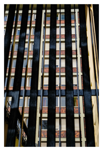 Foto im Hochformat. Blick durch ein Tor mit langen schwarzen Streben. Das Tor ist knapp vor der Kamera. Dahinter ist ein hohes Gebäude mit vielen Fenstern. Es sind immer zwei Fenster nebeneinander. Darunter ist die Wand mit braunen Fliesen gekachelt.