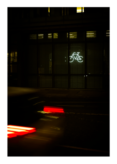 Nighttime photograph showing an illuminated white bicycle symbol on a dark building facade, with red light trails from passing vehicles visible in the foreground, highlighting urban cycling infrastructure.