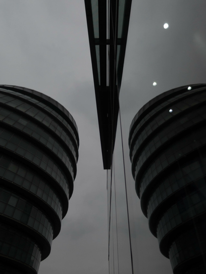Monochrome photo looking up at a part of a round building against a grey sky. On the right is a large window that reflects the building. An sliver of an architectural feature above the window cuts in from the top and is also reflected, a stiletto blade between two opponents.