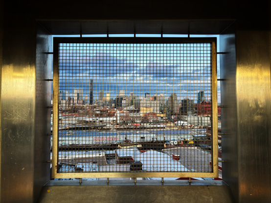 The skyline of downtown Brooklyn at sunset, as seen from the window of the elevated outdoor subway platform at Smith-9th Street. The window screen is a wire grid in a thin “floating”metal frame, which is attached by rods to the deeply recessed window opening. The surrounding metal-plated walls are scuffed and scratched but with a soft golden sheen, illuminated by the sun behind the camera. The window frames a view of low warehouses with graffitied walls, and in the distance, tall buildings under a deepening blue sky with shadowed clouds.