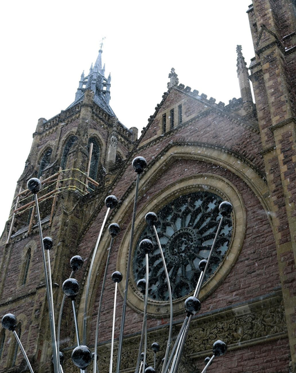 Photograph of a church facade taken from below and slightly at an angle, showing its large rose window and its bell tower, currently under renovation and held up by cables. In the foreground is a modern sculpture composed of silver-colored rods with shiny black spheres at their tips. The church is built of pink and brown bricks, the decorative structure inside the rose window is black, the sky is overcast and white, and snowflakes are falling.

Photographie de la façade d'une église prise du dessous et légèrement de biais, montrant sa grande fenêtre en rosace et son clocher en cours de rénovation et retenu avec des câbles, avec en avant-plan une sculpture moderne composée de tiges de couleur argentée avec des sphères noires luisantes à leur extrémité. L'église est construite de briques roses et brunes, la structure décorative à l'intérieur de la rosace est noire, le ciel est couvert et blanc, et des flocons de neige tombe du ciel.
