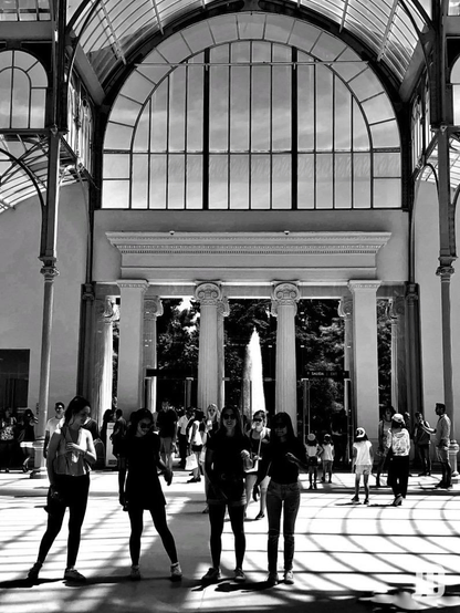 Fotografía en blanco y negro del interior del Palacio de Cristal, capturada desde una perspectiva que resalta su imponente estructura de vidrio y metal.
Destaca el gran arco de medio punto acristalado y las columnas de estilo jónico que enmarcan la salida principal.
El suelo muestra un patrón geométrico de sombras proyectadas por la estructura metálica del techo, creando un contraste visual muy marcado.
El interior está concurrido por visitantes, y a través de la puerta se distingue una fuente de agua en el exterior, rodeada de vegetación.