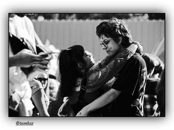 A black and white street photograph captures two young women in a close embrace, gazing into each other's eyes with tenderness and focus. The shorter one on the left has long hair and wears a polka-dotted top, her arms wrapped around the neck of the taller woman who has short hair, glasses and a dark shirt. Their faces are close, their expressions filled with affection. They stand among a crowd, with people smiling and holding up phones. In the background a metal fence is visible. The soft light and grainy texture of the image highlight the intimacy of this moment amid the urban bustle.