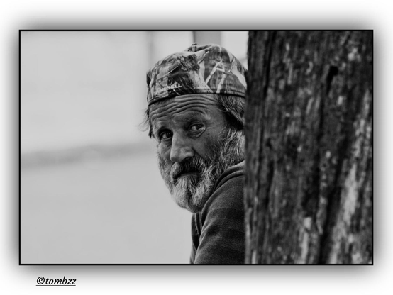 A black and white street portrait shows an older man peeking from behind a tree trunk, glancing over his shoulder as if caught off guard. He wears a sweatshirt and a cap with lettering, slightly askew on his head. His forehead is furrowed, and his gray beard and tousled hair add to the impression of a moment interrupted. His gaze meets the camera with surprise and intensity. The background is softly blurred, drawing attention to the texture of the tree bark and the expression on his face. The image carries a quiet tension, framed in grainy tones that emphasize the fleeting intimacy of the scene.