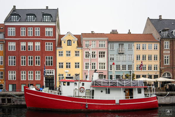 Color photo of a red-hulled boat with a white superstructure moored in front of a row of old multistory houses with differently colored facades of red and yellow and pink and blue.
