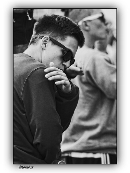 A black and white street photograph shows a young man in the foreground caught mid-gesture as he brushes something off his shoulder. He wears a dark sweatshirt and sunglasses with thick, dark frames. His face is partially obscured by his raised arm, adding a sense of motion and spontaneity. In the background, slightly out of focus, stands another young man in a lighter sweatshirt and sunglasses with pale frames. Around them, other people are visible, forming a lively street scene. The soft light and grainy texture of the image emphasize its candid, documentary character.