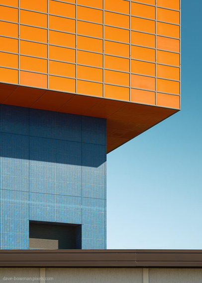 A semi-abstract photograph of the exterior of Osaka Aquarium Kaiyukan, where large, bold panels of blue and orange meet in sharp contrast. The building has clean, precise edges and repeating lines that give it a structured, orderly rhythm. Against the open sky, the composition emphasises the balance of shapes and colours, creating a striking visual effect that blends architectural design with abstract form.