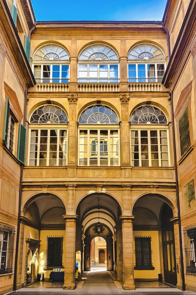 The photo is ofcourtyard with a three-story building facade under a bright blue sky. The building has three stories visible, each with distinct architectural features and windows. The ground level has arched passageways supported by columns, creating a corridor that leads out onto the street. The second story has large, arched windows with white frames and intricate geometric patterns within the arched part. The third story mirrors the second in window design but reflect blue sky and white clouds.
