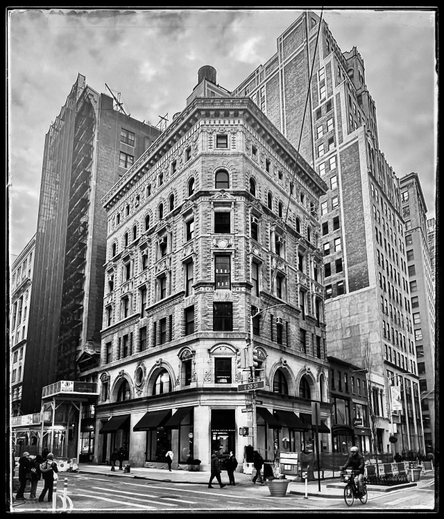 A black and white photo taken from the middle of an intersection towards a corner building. The corner of the building is flat with a storefront on the bottom and six windows going straight up to a decorated balustrade on the roof. It looks like a triangle with the tip cut off. The windows on the second floor over the awnings are large rounded windows. The rest of the building has small windows and an intricate patterned brick work. On the left is a taller building covered in scaffolding. On the right and behind it is a newer and much taller building. In the foreground are people crossing the street and someone on a bicycle. The sky is very cloudy. The coloring and frame around the photo make it feel much older than it is  