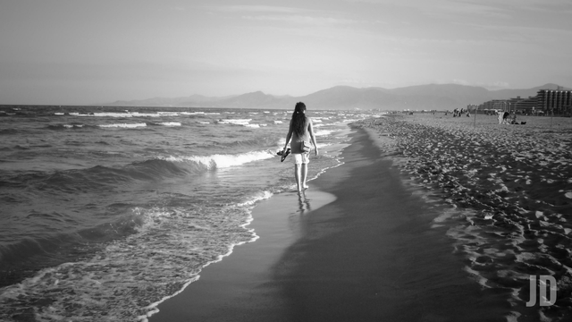 Fotografía en blanco y negro que captura una escena serena en una playa.
Se observa a una persona caminando de espaldas por la orilla, justo donde el agua se encuentra con la arena. Lleva ropa ligera y parece sostener su calzado en la mano.
El mar ocupa el lado izquierdo con pequeñas olas rompiendo suavemente. A la derecha se extiende una amplia playa de arena.
En la distancia se distinguen siluetas de montañas bajo un cielo despejado, y hacia el extremo derecho se aprecian algunas edificaciones costeras y otras personas a lo lejos.
La imagen tiene una perspectiva profunda que guía la mirada hacia el horizonte, transmitiendo una sensación de tranquilidad y soledad contemplativa.