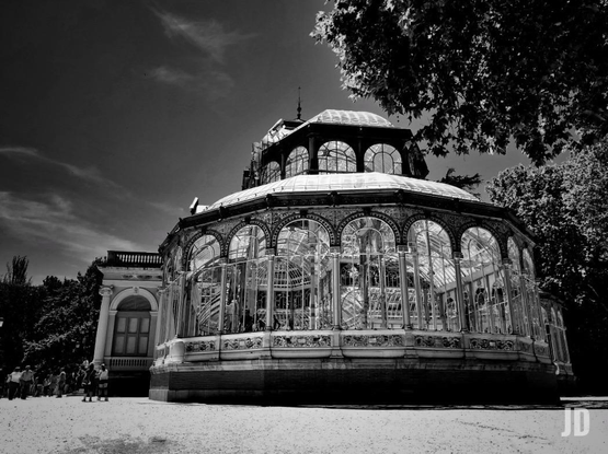 Fotografía artística en blanco y negro que muestra una perspectiva lateral y contrapicada del Palacio de Cristal en el Parque del Retiro.
Se aprecia perfectamente el esqueleto de hierro fundido y las grandes vidrieras que forman las paredes curvas del edificio.
En la parte inferior de los ventanales se distinguen los frisos decorativos de cerámica que bordean toda la estructura.
El uso del blanco y negro resalta el brillo del cristal bajo la luz del sol y crea un contraste dramático con el cielo oscuro y el follaje de los árboles que enmarcan la composición.
La presencia de figuras humanas a la izquierda de la imagen permite apreciar la gran escala y altura del palacio.
Es una toma muy elegante que captura la esencia atemporal del monumento.