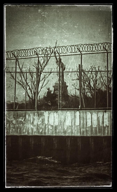 The Statue of Liberty stands in the distance, seen through a chain link fence topped with coils of barbed wire in the foreground, on a weathered concrete pier rising over dark choppy waters. The perspective is such that the statue appears to be imprisoned behind the fence, a detainee in a concentration camp. The image has been edited to look like a faded and heavily scratched monochrome print with a distressed black border — a bleak document of an atrocity straight out of the history books.

I used to consider myself lucky to live within walking distance of a view of my city’s (and perhaps this country’s) most iconic symbol, but now it’s a bitter sight: a reminder of ideals betrayed, or lies that were never true to begin with. A nation as hollow as the statue itself. There’s still much to love about this place, but it has more to do with good people on the ground, in their unnoticed corners and unremarked lives, bringing joy and creating art and fighting the dark. My faith’s in them. I’m done venerating institutions and national mythologies.