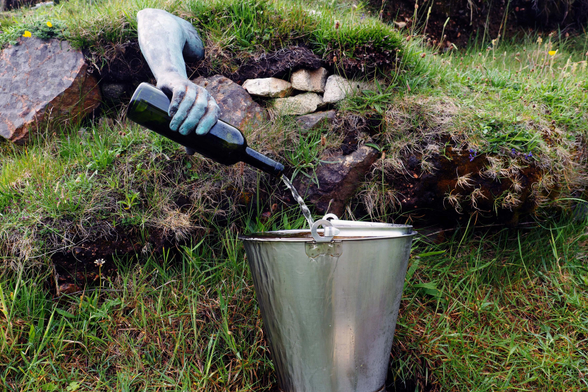 Grünes Gras, ein paar Steine am Wegesrand, eine kleine Quelle: Ein Arm ragt aus einem Hügel hervor, und eine Hand hält eine Flasche, aus der Wasser in einen Zinkeimer fließt – gefunden 2017 auf den Hebriden, Lewis.

Green grass, a few stones by the side of the path, a small spring: an arm protrudes from a hill and a hand holds a bottle from which water flows into a zinc bucket – found in 2017 on the Hebrides, Lewis.