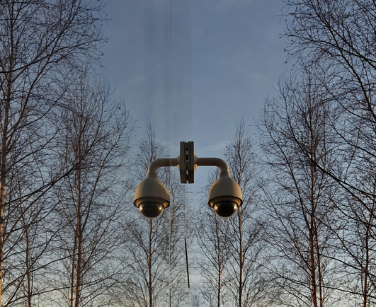 A photo taken along a reflective wall, with some leafless trees in front of a pale blue evening sky. A surveillance camera attached to the wall seems to be floating in the air.