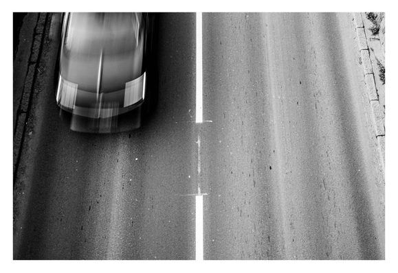 Schwarz-weiß Foto im Querformat. Blick von oben auf eine einspurige Straße. Die rechte Spur ist leer, auf der linken Spur kommt gerade von oben ein dunkles Auto ins Bild. Durch eine längere Belichtungszeit hat dieses Bewegungsunschärfe. Der Mittelstreifen der Fahrbahn ist genau mittig im Bild.