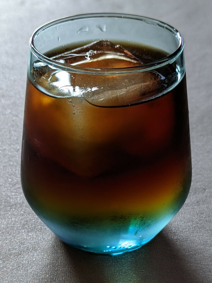 Photo of a blue glass on a grey background. In the glass is a large ice cube and coffee that fades to clear water at the bottom.