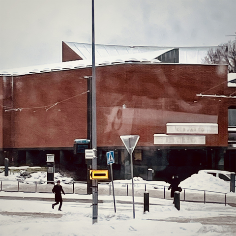 The same snowy street scene without the superimposed cat face, showing a modern red-brick building partially obscured by snow, with two people walking nearby.