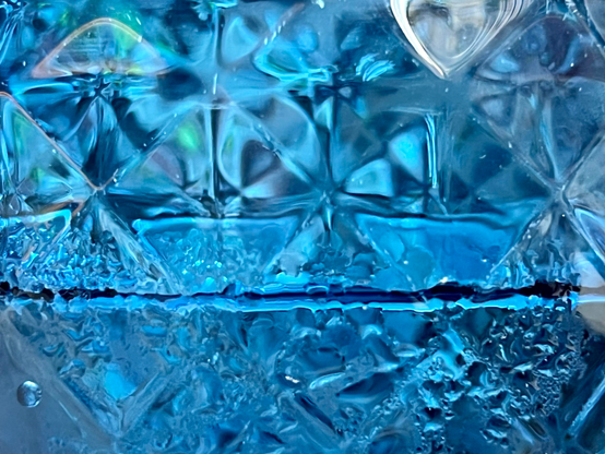 Abstract closeup side view of an ice-blue glass tumbler half-full of water, with an embossed diamond-shape recurring pattern in the glass, which look a bit like four-leaf clovers and catch the light in all sorts of shades of oil-slick. The water level is about halfway up the frame, with some bubbles visible below the surface, and some condensation droplets on the outside of the glass. 