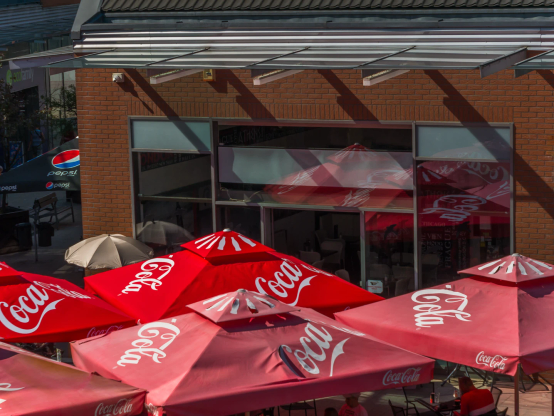 Black von oben auf den Eingang und den Außenbereich eines Cafés.
Kräftig rote und auch ausgeblichenere Coca Cola Schirme (die sich auch in der Glasfront spiegeln) beherrschen die Szene.
Links lugt ein vorwitziger Pepsischirm ums Eck.