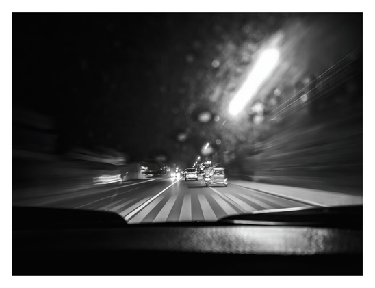 A moody, black-and-white photograph taken from inside a moving vehicle at night. The image conveys a strong sense of speed through motion blur, particularly visible in the streaked streetlights and the passing surroundings. The view looks out over a dark dashboard onto a road with distinct striped markings, following traffic ahead. - Google Gemini 3 Pro Preview