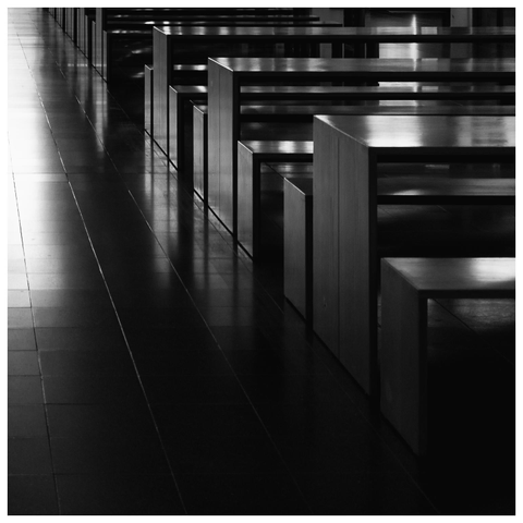 Schwarzweißfoto mit einer Reihe identischer, kantiger Tische oder Bänke, perspektivisch nach hinten versetzt, auf einem glänzenden Boden; starke Linien und Kontraste, ruhige, leere Atmosphäre.