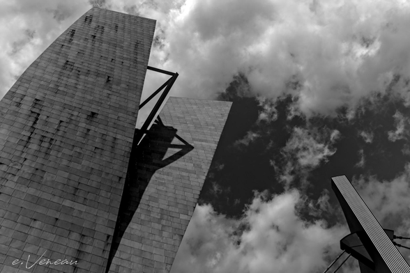 From a low angle, the towers of the museum converge towards a cloudy sky. 2 curved towers in profile, one of which shades the other. From the front with vertical lines a pillar of the bridge.