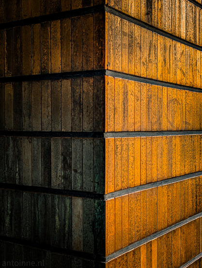 A vertical line runs down the center of the frame, marking the sharp 90-degree corner of a pillar. The right side is bathed in a warm, golden-orange glow from the low sun, while the left side falls into deep, moody shadows.

The walls are composed of narrow, vertical rectangular tiles. These are separated by thick, dark horizontal recessed bands, creating a rhythmic, grid-like pattern.

The image is dominated by deep ambers, burnt oranges, and earthy browns, contrasted against heavy blacks and dark charcoal tones in the recessed areas.