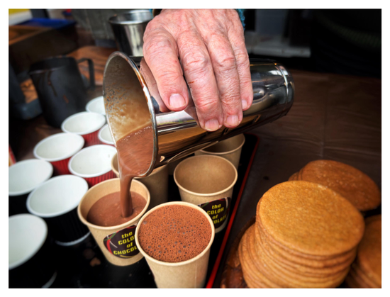Closeup of an older person’s hand pouring hot chocolate from a steel tumbler into small paper cups arranged in rows on a wooden counter. The cups to the side are red or black with white interiors, while the cups at the center of the image are brown and display circular stickers that say “The Color of Chocolate.” To the right of the cups are stacks of thin brown cookies. In the background is a blurred pitcher and other kitchen equipment.