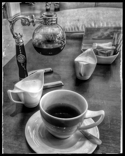 Im Vordergrund eine schlichte weiße Tasse,mit Kaffee, dahinter ein Milchkänbchen, ein Kaffeebrühsiphon mit einer Glaskugel als Brühbehälter, ein Gefäß mit Zuckertütchen, und ein Kissen, das auf einem Sessel hinter dem Tisch hervorlugt