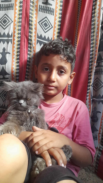 Young boy (Saji) with curly brown hair wearing a pink shirt, cradling a fluffy gray cat with a small silver bell collar, sitting on his lap; the background features patterned fabric.