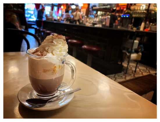 A cozy scene at an ice cream parlor. In the foreground is a stout glass mug of hot chocolate on a saucer with a stirring spoon, atop a table with marble-like swirling. The chocolate is a deeper brown on the bottom with a lighter layer on top where it mixes with cream. Rising above the brim is a toasted marshmallow topped by a mountain of whipped cream, all dusted with flakes of grated chocolate. Blurred in the background are barstools in front of a long counter, behind which are workers and parlor equipment. Daylight shines through the windows at the far end.