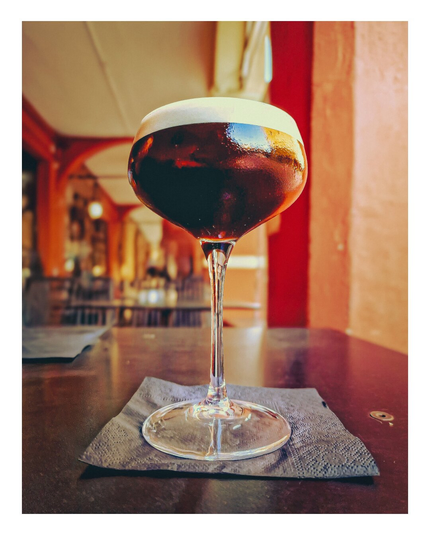 A photograph of a single, large, clear wine or cocktail glass (I don't know my glasses) filled with dark, caffè shakerato, standing on a gray/blue napkin on a dark table. The background shows a warmly lit, rustic bar with red walls, beams, and blurred bar stools.